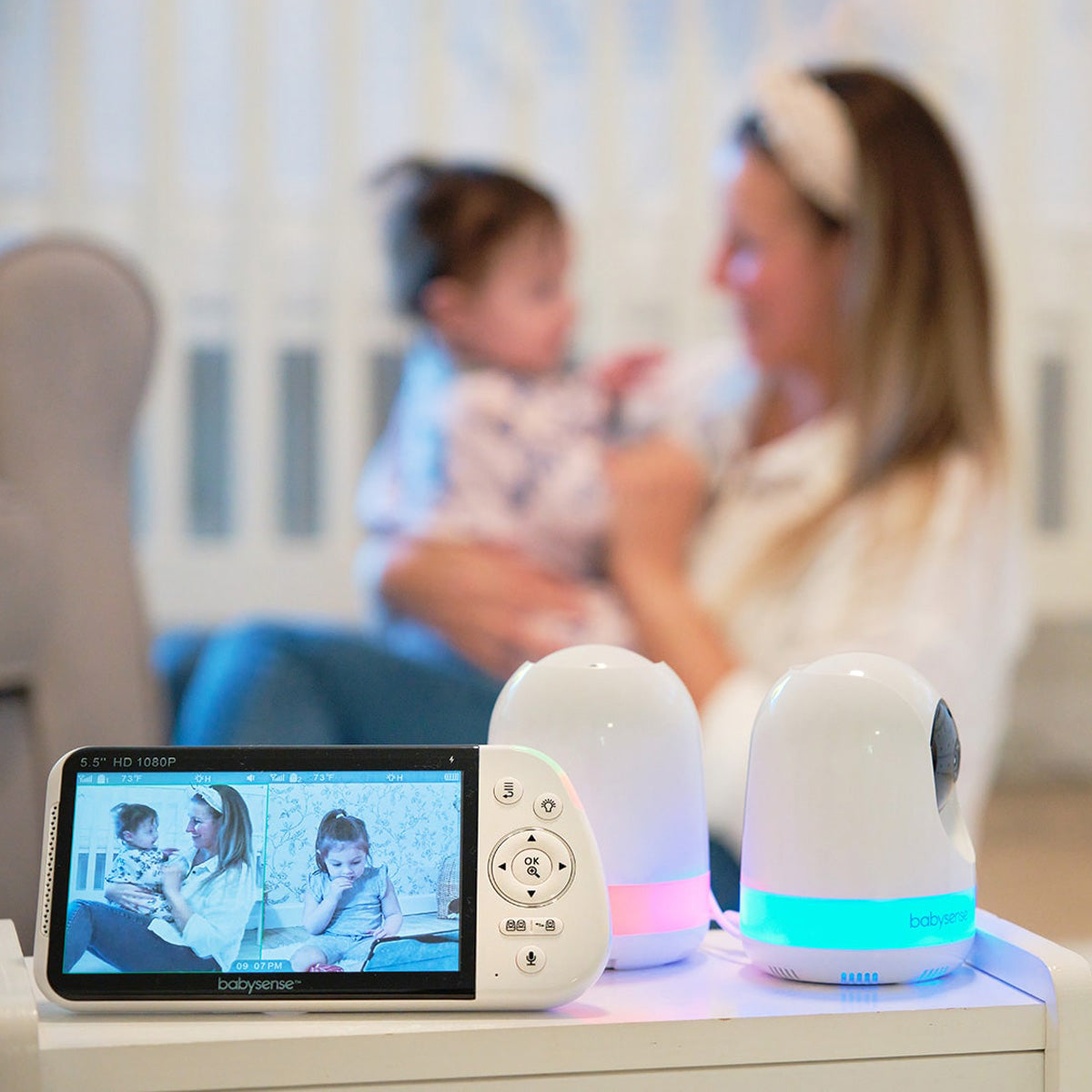 A white Babysense handheld monitor displays two different nurseries, showing a baby and a toddler, next to two Babysense cameras with glowing bases, making them the best baby white noise machine