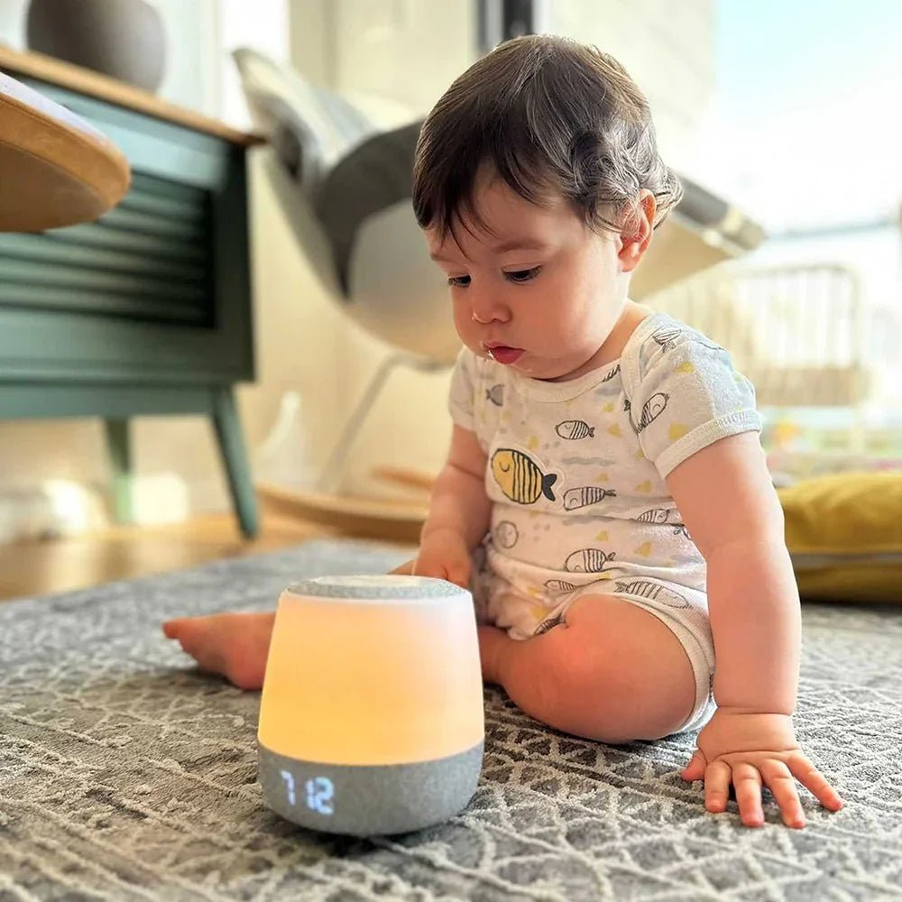 A curious baby wearing a fish-patterned onesie sits on a gray patterned rug, looking down at the illuminated white and gray Babysense night light sound machine showing “7:12” on its base, providing essential baby safety products for home