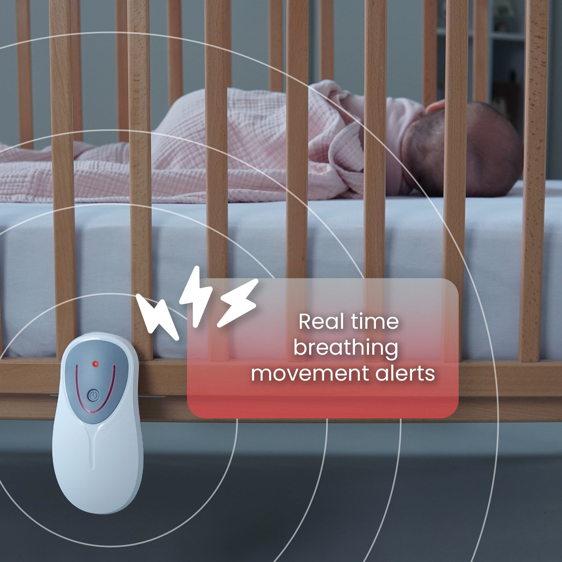 A sleepy baby dressed in pink rests covered by a light blanket in a wooden crib, alongside a white Babysense breathing-motion monitor providing real-time movement alerts, demonstrating the Babysense Ultimate Safety Bundle